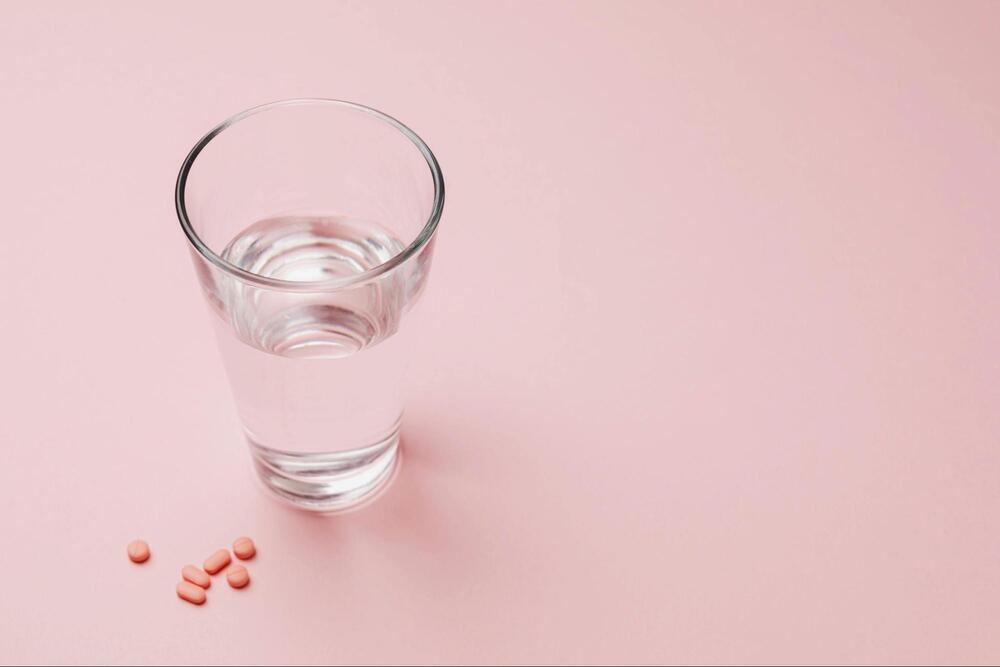 A glass of water and pink pills on a light pink background