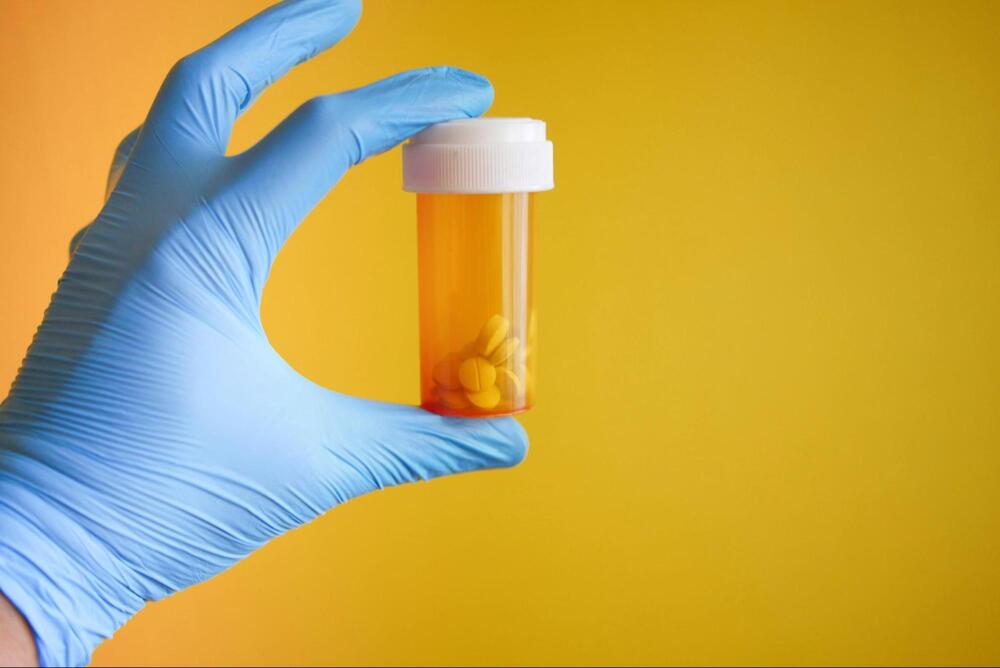 A gloved hand holding a bottle of medicine against an orange background